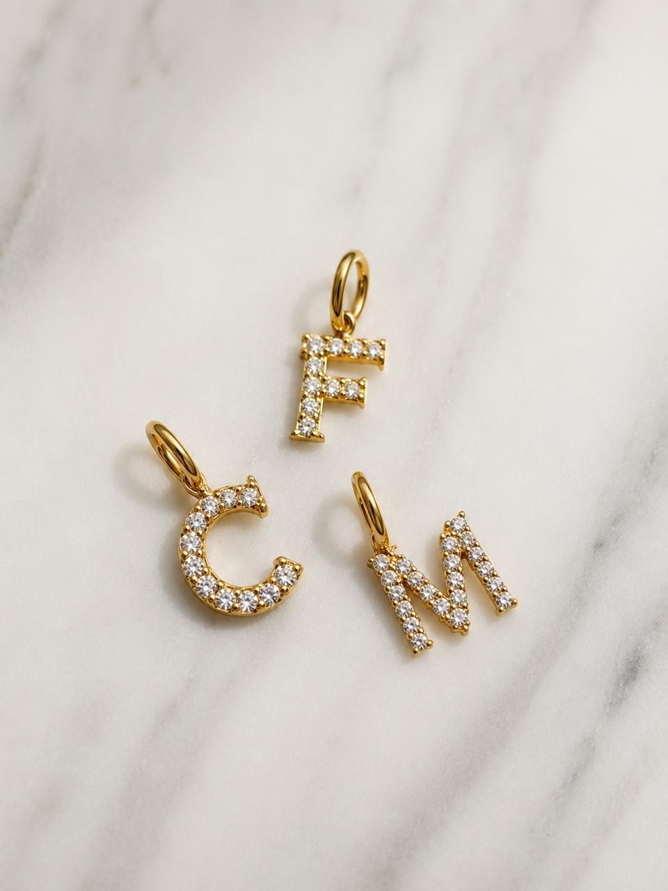 Dos dijes dorados con forma de letras y pequeñas piedras claras sobre una superficie blanca.